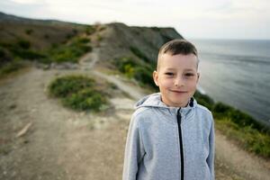 Porträt von ein Junge Stehen auf das Kante von ein Cliff in der Nähe von Meer. foto