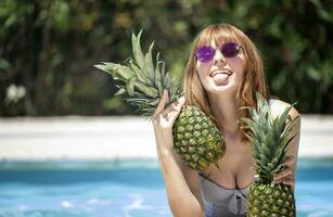 kaukasisch Mädchen mit Sonnenbrille halten zwei Ananas im ein Schwimmbad. foto