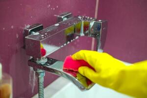 Rührgerät mit Reinigung Agent. ein Hand im ein Gummi Handschuh reinigt das Wasserhahn mit ein Schwamm im das Badezimmer. foto