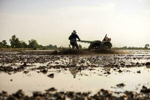 niedrig Winkel Sicht, Farmer ist Fahren hinterherlaufen Traktor zu Pflug wassergefüllt schlammig Boden. foto