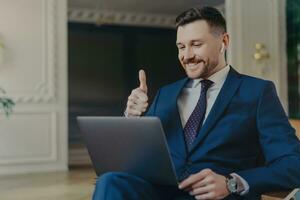 glücklich Geschäft Fachmann im elegant Anzug, zeigen Befriedigung mit ein Daumen hoch Geste während ein Video Konferenz mit Boss oder Kollegen auf ein Laptop. Arbeiten im ein modern, lichtdurchflutet Büro. foto