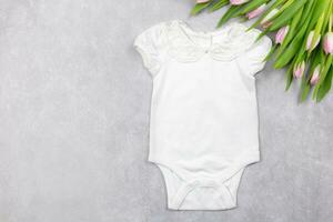 Weiß Baby Mädchen Bodysuit Attrappe, Lehrmodell, Simulation eben legen mit Rosa Tulpen Blumen auf das grau Beton Hintergrund. Design Strampler Vorlage, drucken Präsentation spotten hoch. oben Sicht. foto