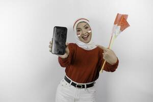 ein Porträt von ein lächelnd asiatisch Muslim Frau tragen rot oben und Weiß Hijab und zeigen ihr Telefon Bildschirm, isoliert durch Weiß Hintergrund. Indonesiens Unabhängigkeit Tag Konzept foto