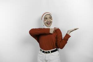 ein glücklich asiatisch Muslim Frau tragen rot oben und Weiß Kopftuch, und zeigen Kopieren Raum neben ihr, isoliert durch Weiß Hintergrund. Indonesiens Unabhängigkeit Tag foto