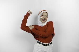 aufgeregt asiatisch Muslim Frau tragen ein rot oben und Weiß Hijab zeigen stark Geste durch Heben ihr Waffen und Muskeln lächelnd stolz. Indonesiens Unabhängigkeit Tag Konzept. foto