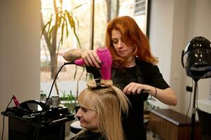 modisch Friseur mit tätowiert Waffen mit ein runden Kamm und Haar Trockner richtet sich auf blond Haar zu ein Klient von ein Schönheit Salon, durchführen ihr ein stilvoll Frisur. Schönheit Industrie Konzept foto