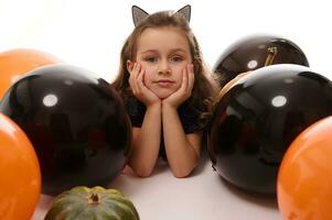 schön Kind Mädchen mit Band mit Katze Ohren sieht aus beim Kamera Lügen Nieder Nächster zu Kürbis, farbig schwarz und Orange Luftballons auf ein Weiß Hintergrund mit Kopieren Raum. Halloween Konzept foto