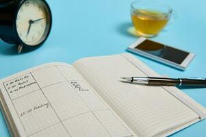 ein öffnen Agenda mit Zeitplan, Smartphone, Tinte Stift, Glas Tasse von Tee und schwarz Alarm Uhr Lüge auf Blau Oberfläche. Farbe Hintergrund mit Kopieren Raum . Zeit Management, Termin, Zeitplan Konzept foto