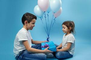 ein wenig Junge gibt ein Geschenk zu ein wenig Mädchen Sitzung im Vorderseite von ihm gegen ein Hintergrund von farbig Luftballons auf ein Blau Oberfläche foto