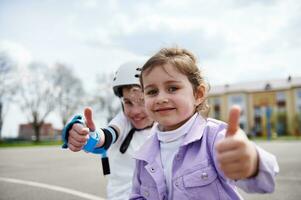 Gesicht Porträt von bezaubernd Kinder, Junge im Skateboard Helm und Mädchen Sitzung Nächster zu jeder andere und zeigen Daumen oben foto