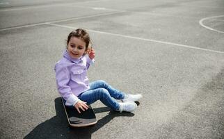 süß wenig Mädchen Sitzung auf ein Skateboard auf das Spielplatz auf ein schön sonnig Tag foto