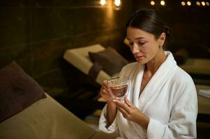 heiter schön elegant Mitte alt europäisch Frau im Weiß Bademantel, mit ein Tasse von gesund Kräuter- Tee im Hände, genießen ayurvedisch Schönheit Behandlung, Sitzung durch Whirlpool im Luxus Wellness Spa Center foto