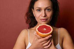 Nahansicht. attraktiv Mitte alt ziemlich Frau mit Hälfte von frisch saftig rot Grapefruit, suchen beim Kamera, isoliert auf Koralle Hintergrund. Gesundheitspflege, Medizin, Diät und gesund Essen, Lebensstil Konzept foto