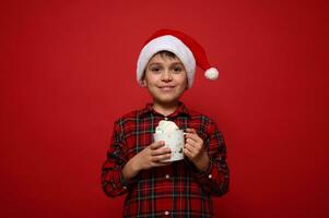 Porträt von tolle preteen süß Junge, bezaubernd Kind im Santa hatand Plaid Hemd halten ein Becher it heiß Schokolade mit Marshmallows, lächelt suchen beim Kamera gegen rot Hintergrund mit Kopieren Raum foto