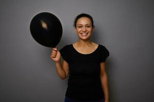 heiter afrikanisch amerikanisch Frau gekleidet im schwarz lächelt suchen beim Kamera und posiert gegen grau Hintergrund mit ein schwarz farbig Luft Ballon im Hand. schwarz Freitag Konzept mit Kopieren Anzeige Raum foto