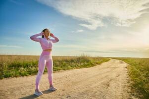 in voller Länge Sport Frau im Kopfhörer Stehen auf das Steppe Straße bereit zu Joggen im Natur im das Licht von Sonnenuntergang foto