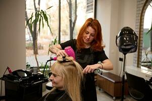 modisch schön rothaarig weiblich Friseur mit ein runden Kamm und Haar Trockner richtet sich auf blond Haar zu ein Klient von ein Schönheit Salon, durchführen ihr ein stilvoll Frisur. Schönheit Industrie Konzept foto