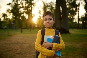 zuversichtlich Porträt von ein glücklich gut aussehend Schule Kind Junge 9 Jahre alt mit Rucksack halten Arbeitsmappen und Bleistift Fall im Hände und süß lächelnd posieren zu Kamera gegen Natur Hintergrund beim Sonnenuntergang foto
