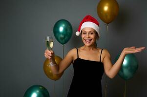 schön Frau im Santa Hut freut sich und hat Spaß beim Weihnachten Party hält ein Champagner Flöte und Kopieren Raum auf ihr Hand Palme hoch, isoliert auf grau Hintergrund mit glänzend Gold und Grün Luft Luftballons foto