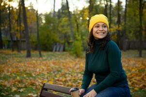 heiter Erfreut Frau im hell bunt warm gemütlich Kleider ruhen auf ein Park Bank mit ein Karton Kaffee Tasse auf das Hintergrund von herbstlich Ahorn und Eiche Bäume, süß lächelt suchen beiseite foto