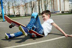 wenig Junge rutschte aus und fiel aus das Skateboard auf das Asphalt von das Spielplatz foto