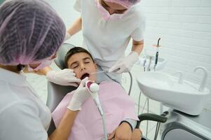 mutig Schule Junge Sitzung auf Zahnarzt Stuhl Empfang medizinisch Behandlung von seine Oral Hohlraum durch Kinderarzt Zahnarzt und seine Assistent im modern Zahnheilkunde Klinik foto