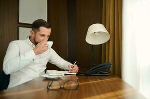 Porträt von ein gut aussehend attraktiv Geschäftsmann, Unternehmer, Journalist, Schriftsteller halten Silber Stift und nehmen Anmerkungen im Notizbuch während Trinken köstlich frisch gebraut Kaffee im seine Büro foto