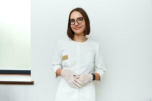 zuversichtlich Porträt von ein jung Arzt im Weiß medizinisch Labor Mantel und schützend Handschuhe suchen beim Kamera während posieren gegen Weiß Mauer im Kosmetologie Klinik foto