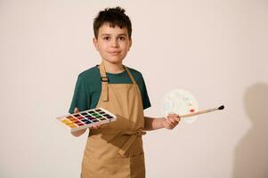 kaukasisch gut aussehend talentiert Teenager Junge halten Palette mit Aquarelle und Farbe Bürste, lächelt beim Kamera. Kunst Klasse foto