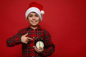 Hüftumfang Porträt von bezaubernd Junge mit ein schön zahnig lächeln, tragen ein Santa Hut und zeigen das Zeit auf das wählen von ein schwarz Alarm Uhr im seine Hände. es ist Mitternacht. fröhlich Weihnachten Konzept foto