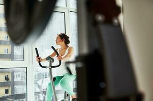 schön Frau sieht aus gegenüber das Fenster während ausüben auf ein Übung Fahrrad. gesund Gewohnheiten Konzept foto