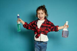 schön wenig Mädchen posieren auf ein Blau Hintergrund mit Waschmittel Sprays im ihr ausgestreckt Waffen foto