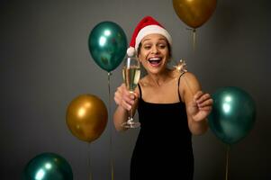 attraktiv Frau mit schön Lächeln im schwarz Abend Kleid und Santa Hut genießt Weihnachten Party, hält Champagner Flöte und Bengalen Beleuchtung, freut sich auf grau Hintergrund mit festlich funkeln Luft Bälle foto