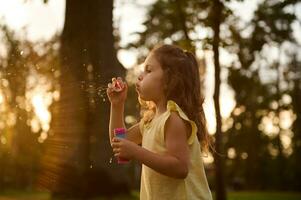 schön Herrlich süß bezaubernd wenig Kind Mädchen weht Seife Luftblasen beim Sonnenuntergang. sorglos Kindheit, draußen Freizeit Aktivitäten. Baby Mädchen spielen Spiele im das Natur foto