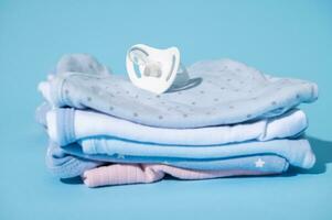 ein Baby Tröster auf ein Stapel von sauber gebügelt Neugeborene Bodys, isoliert Über Blau Pastell- Hintergrund. erwarten ein Baby foto