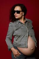 attraktiv lockig multiethnisch schwanger Frau tragen modisch Sonnenbrille und beiläufig Denim Kleidung, isoliert rot Hintergrund foto