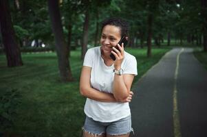 schön multiethnisch jung Frau reden auf Handy, Mobiltelefon Telefon während Gehen entlang das Gasse von ein Stadt Park auf ein Sommer- Tag foto