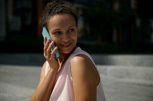 Nahansicht Porträt von ein schön afrikanisch amerikanisch Frau reden auf Handy, Mobiltelefon Telefon, suchen weg, posieren zu Kamera auf städtisch Gebäude Hintergrund foto