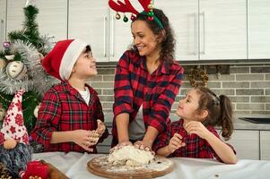 glücklich Mutter mit Geweih Band auf Kopf bereitet vor, knetet Teig mit Rosinen und Nüsse auf ein hölzern Planke, lächelt reden mit ihr bezaubernd Kinder, genießt Ausgaben Zeit zusammen während Weihnachten Ferien foto