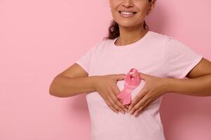abgeschnitten Porträt auf farbig Hintergrund von ein lächelnd Frau im Rosa T-Shirt, Putten Hände auf Truhe im gestalten von Herz mit ein Rosa Satin- Band im das Center. Welt Krebs Bewusstsein Tag, Kampf Krebs foto