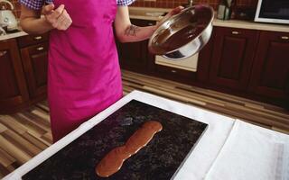 Konditor Chocolatier tragen Rosa Schürze Gießen geschmolzen Schokolade auf Marmor Oberfläche auf das Hintergrund von hölzern Küche foto