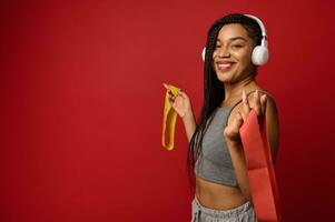 Seite Porträt von ein lächelnd afrikanisch sportlich Frau mit afro Dreadlocks und kabellos Kopfhörer halten bunt elastisch Fitness Bands im Hände, posieren gegen ein rot Hintergrund mit Kopieren Raum foto