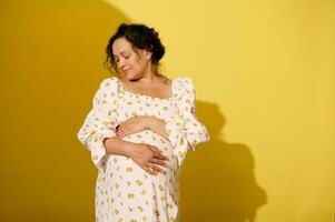 herrlich schwanger Frau sanft streicheln und umarmen ihr groß Bauch, isoliert Über Gelb Studio Hintergrund foto