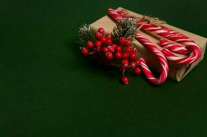 Weihnachten Geschenk im Kunst Verpackung Geschenk Papier dekoriert mit Stechpalme und Süss zuckerhaltig gestreift Weiß und rot Lutscher Süßigkeiten Stöcke gelegt aus im das Ecke von Kopieren Raum auf dunkel Grün Hintergrund foto