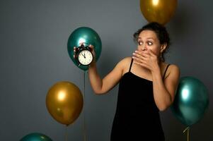 spanisch ziemlich Frau tragen ein Abend schwarz Kleid, Abdeckungen ihr Mund, überrascht durch das Zeit im das Alarm Uhr, es ist Mitternacht. isoliert Über grau Hintergrund mit golden Grün Luft Luftballons Kopieren Raum foto