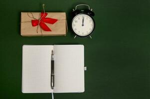 eben legen Komposition mit Stift auf geöffnet Notizblock mit leeren leer Blätter, Alarm Uhr und ein Geschenk im Kunst Verpackung Papier mit gebunden Bogen und rot Band auf dunkel Grün Hintergrund mit Kopieren Raum zum Anzeige foto