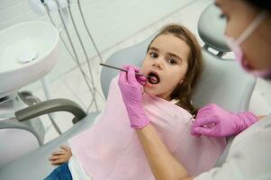 Overhead Aussicht von süß Baby Mädchen beim regulär Dental Untersuchung im Weiß Dental Klinik mit modern Ausrüstung. pädiatrisch Zahnarzt Prüfung Kinder Zähne mit steril rostfrei Stahl Dental Instrumente. foto