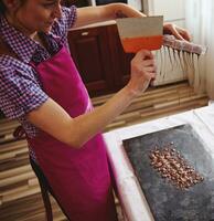 ein Chocolatier mit Kuchen Schaber zu entfernen Überschuss Schokolade von Formen in ein Marmor Oberfläche zu machen Süßigkeiten Muscheln foto