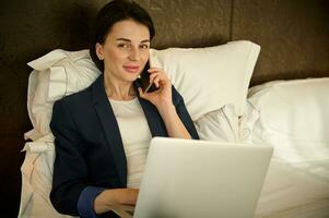 Herrlich wohlhabend Geschäft Frau Herstellung Anruf auf das Telefon und Arbeiten auf Laptop, süß lächelt suchen beim Kamera während Lügen auf das Bett im Hotel Zimmer nach erfolgreich Treffen mit Geschäft Partner foto