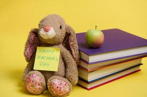 ein Plüsch Spielzeug Hase mit Beschriftung glücklich Lehrer Tag auf Gelb Hinweis Papier, beugte sich vor auf ein schüren von bunt Bücher, Gelb Hintergrund mit Raum zum Text foto